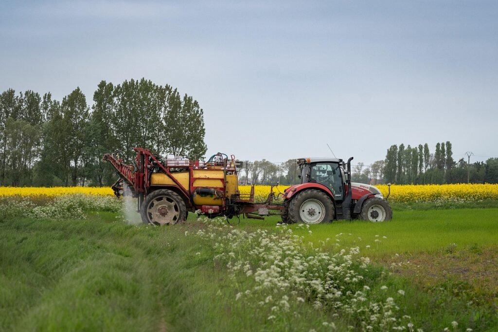 Tractor rojo con pulverizador remolcado tratando un campo, con un cultivo de colza en flor al fondo